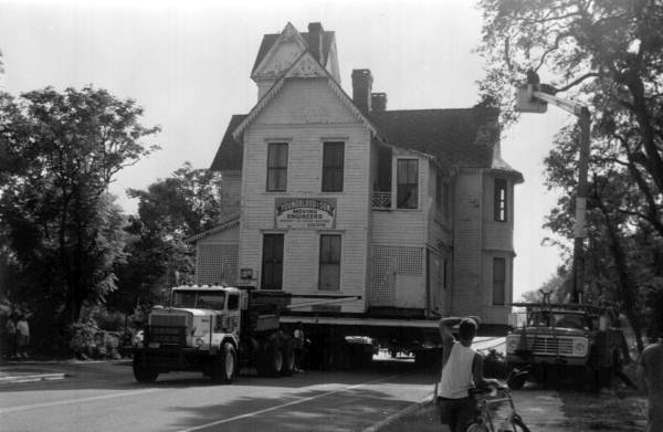A black & white photo of a large flatbed truck carrying a historic Victorian-style home to a new location. A bicyclist looks on in the foreground.